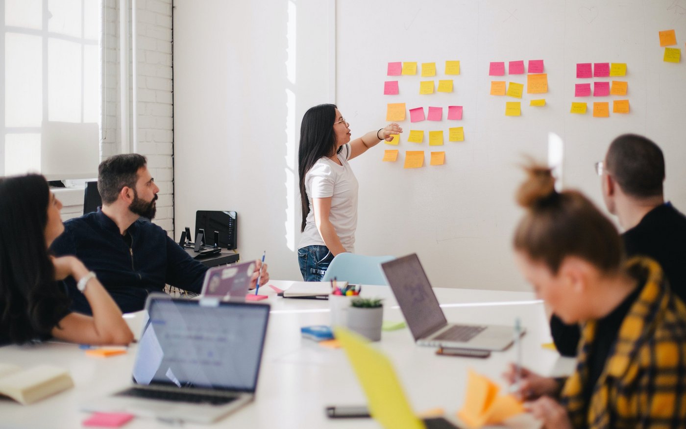 OKR Planning OKR Planning in agilem Setting: Die OKR-Masterin sortiert die Vorschläge mit Haftzetteln an einer Wand, während das Team zuhörend oder arbeitend am Tisch sitzt.