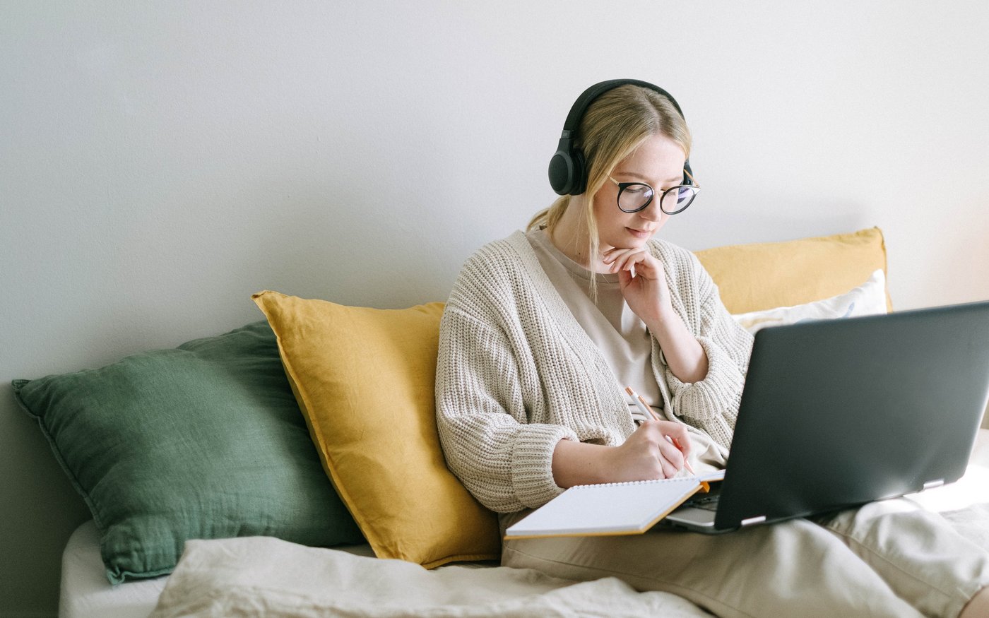 Eine junge Frau mit Brille und Kopfhörern sitzt mit einem geöffneten Laptop auf dem Sofa. Dabei schreibt sie nachdenklich mit einem Stift in ein Notizbuch.