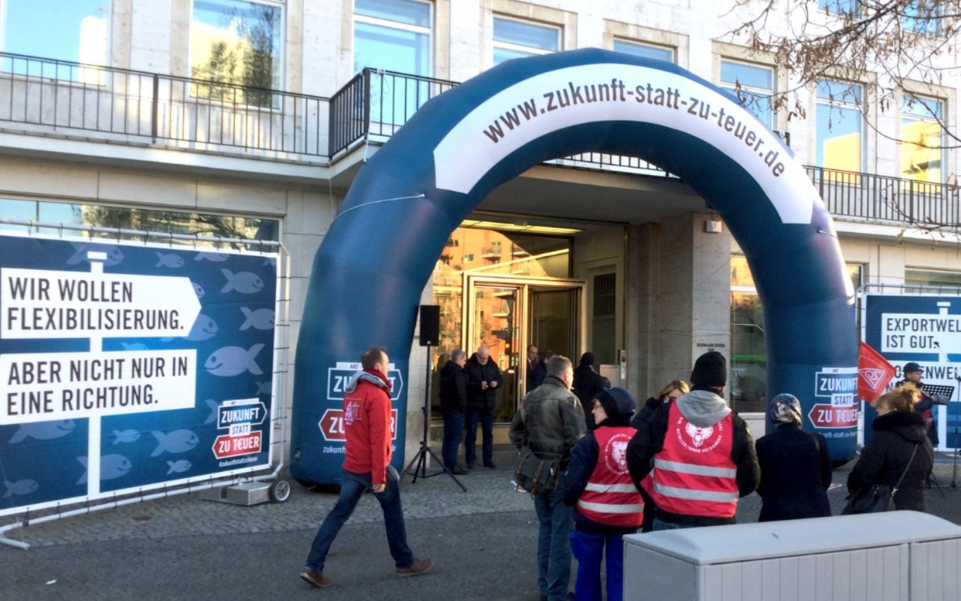 Ein aufblasbarer Bogen mit der Aufschrift 'www.zukunft-statt-zu-teuer.de' am Eingang eines Verhandlungsortes. Links davon ein Plakat mit einem Wegweiser 'Wir wollen Flexibilisierung', sowie einem entgegengesetzten Wegweiser mit der Aufschrift 'Aber nicht nur in eine Richtung'.