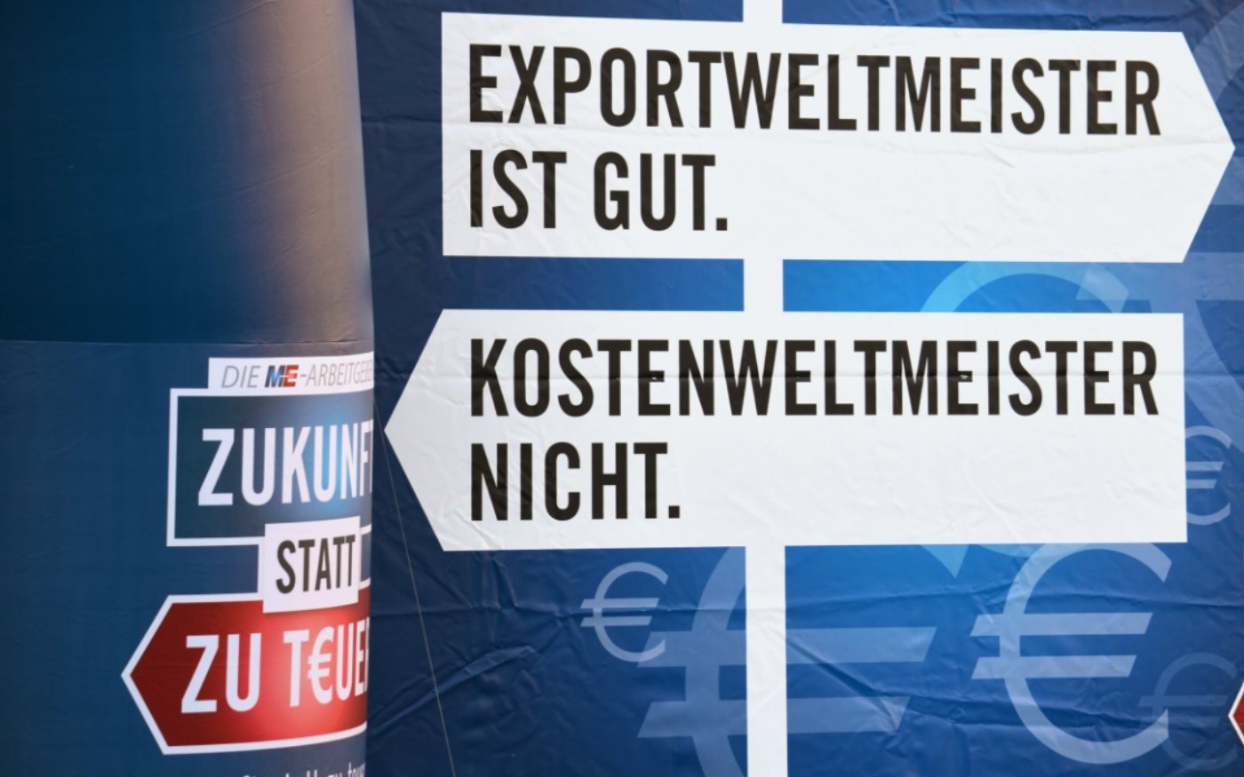 Ein Plakat mit einem Wegweiser mit der Aufschrift 'Exportweltmeister ist gut.', ein weiterer Wegweiser mit der Aufschrift 'Kostenweltmeister nicht.' weist in die entgegengesetzte Richtung.