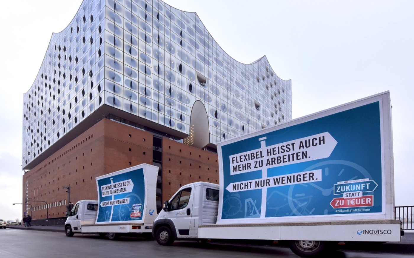 Zwei Fahrzeuge mit Plakaten, darauf ein Wegweiser mit der Aufschrift 'Flexibel heisst auch mehr zu arbeiten.', ein weiterer Wegweiser mit der Aufschrift 'Nicht nur weniger.' weist in die entgegengesetzte Richtung. Im Hintergrund die Elbphilharmonie in Hamburg.