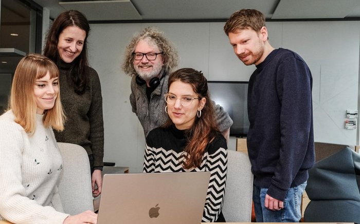 Fünf Personen aus dem Team stehen gemeinsam Um einen Laptop auf dem eine Designerin einen Entwurf zeigt. Alle wirken glücklich.