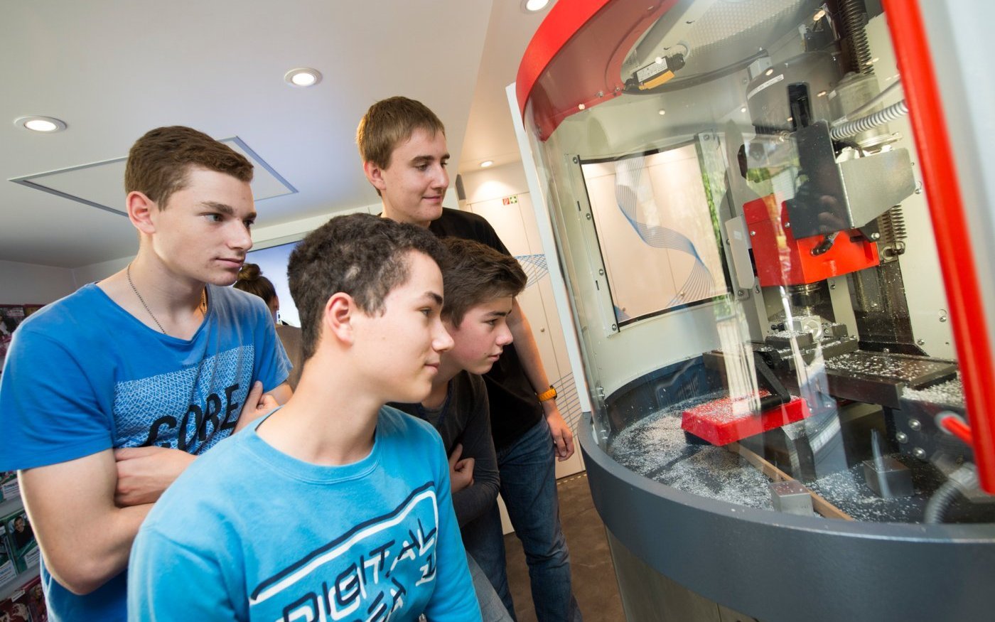 Vier Schüler im Inneren des Trucks beobachten eine CNC-Fräse bei der Arbeit an einem Metallwürfel.