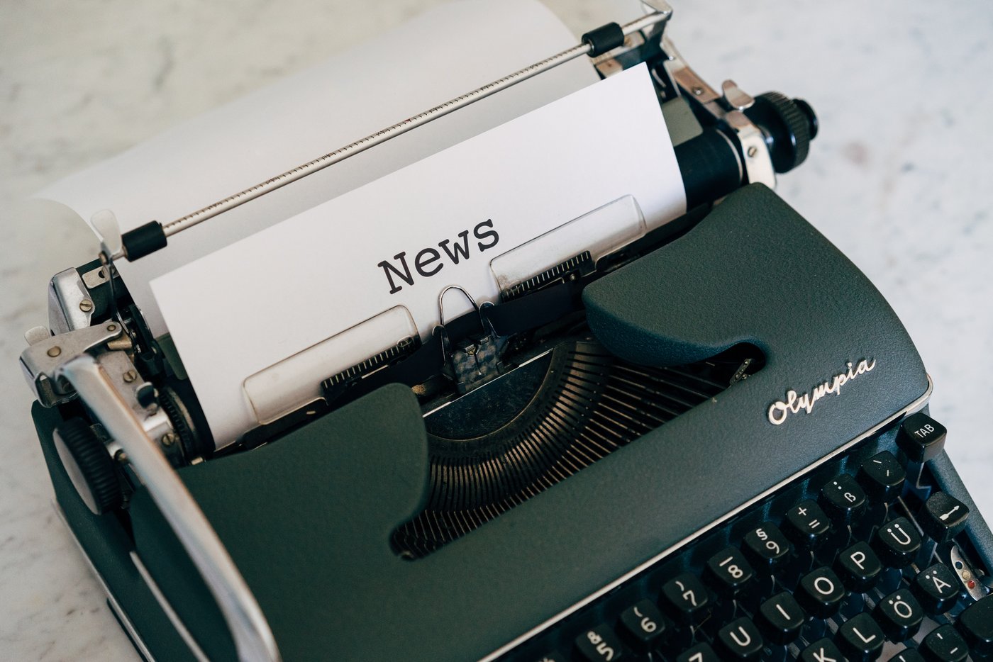 Es wird eine Olympia Schreibmaschine gezeigt, in welcher ein Blatt eingelegt ist, auf welchen die Überschrift 'News' zu lesen ist.