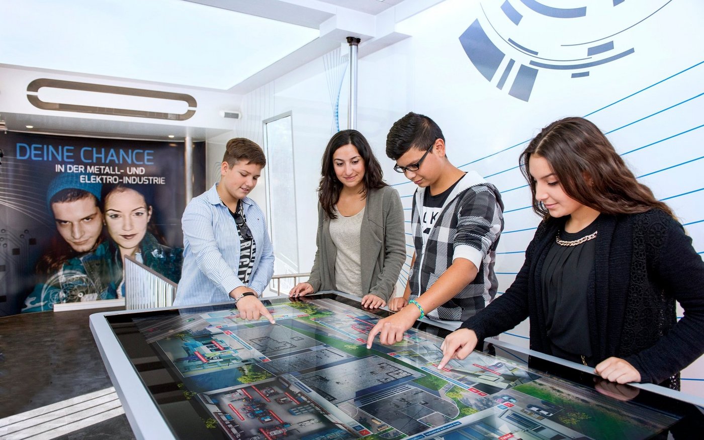 Vier Schüler erkunden an einem Tisch mit integrierten Touchscreen die diversen Ausbildungsberufe in der Metall- und Elektroindustrie.