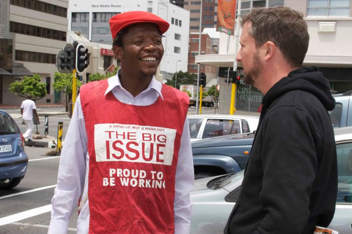 Zwei Männer unterhalten sich an einer Straßenkreuzung. Einer von ihnen trägt eine auffällige rote Weste mit der Aufschrift 'The big issue - Proud to be working'.